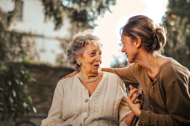 An elderly mother has a conversation with her adult daughter as they hold hands.