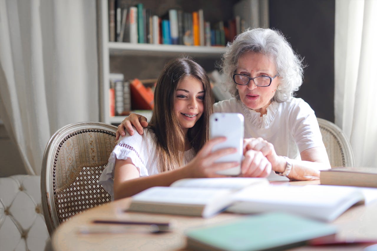 A grandmother in her home sits at a table, looking at a cell phone with a young girl