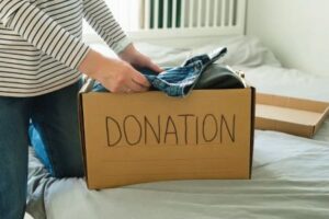 A carboard box with the word "donation," written on it sits on a bed with white sheets. A Caucasian woman's hands can be seen putting a pair of denim pants into the box. She is wearing a white shirt with black stripes.