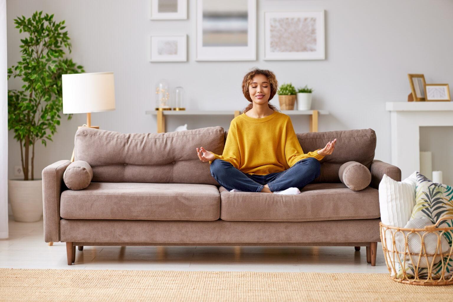 A woman in yellow sits cross-legged on a sofa in a meditative pose. The room around her is clean and organized.