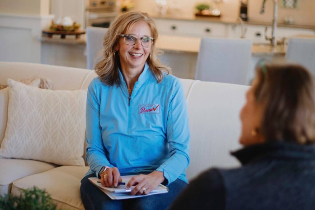 Cheryl, a woman with shoulder length blonde hair sits on a couch talking to a client, who has her back to the camera. Cheryl is wearing a blue shirt with the Consider it Done logo on the breast. She holds a clip board and pen and is smiling in conversation with her client.
